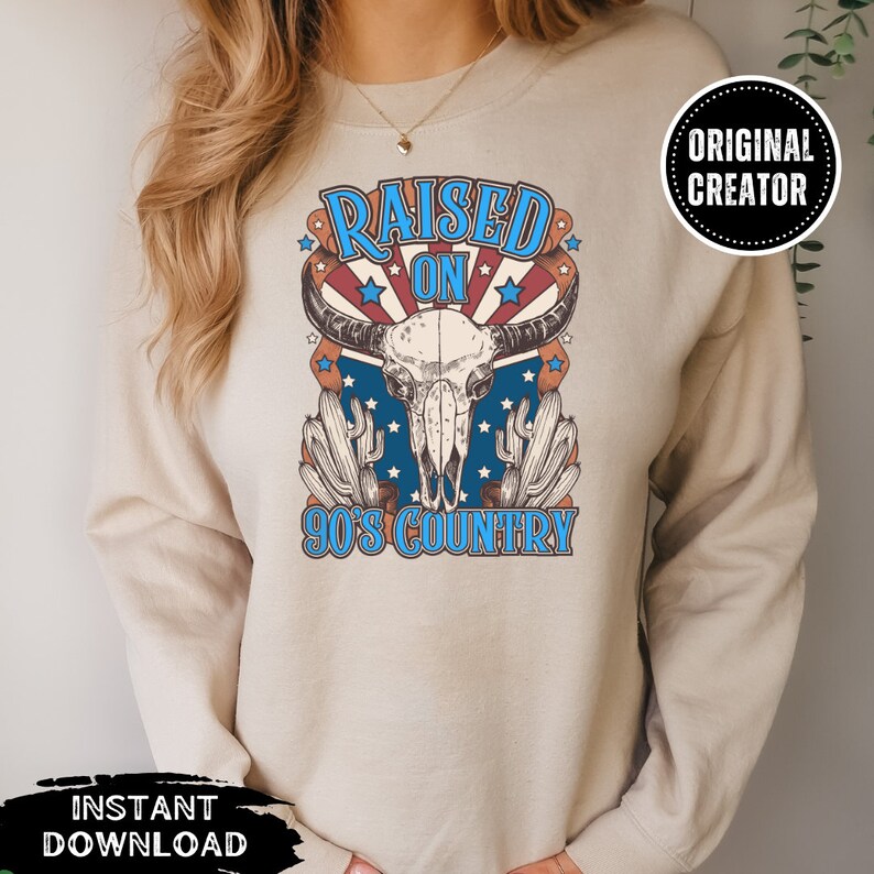 Pu&ograve; includere: Una felpa beige con un design grafico di un teschio di mucca con il testo "Raised on 90's Country" in blu e rosso.
