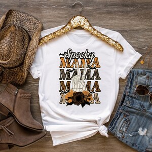 May include: White t-shirt with the text "Spooky Mama" in orange and black lettering. The text is layered with a skeleton hand making a peace sign and a floral design.
