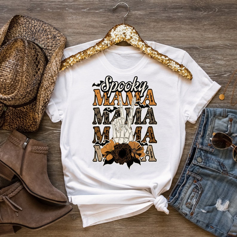 May include: White t-shirt with the text "Spooky Mama" in orange and black lettering. The text is layered with a skeleton hand making a peace sign and a floral design.