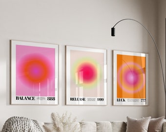 Peut inclure: Trois impressions d'art abstrait encadrées avec un dégradé de couleurs rose, orange et jaune. Les impressions présentent un cercle au centre et les mots "Balance 888", "Release 999" et "Luck 777" en bas. Les impressions sont accrochées à un mur blanc au-dessus d'un canapé blanc avec une couverture et des coussins.