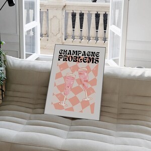 May include: A framed print with the words "CHAMPAGNE PROBLEMS" in black and pink, featuring two champagne glasses on a pink and white checkered background. The print is displayed on a beige sofa near a window.
