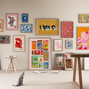 Pode incluir: Uma cole&ccedil;&atilde;o de impress&otilde;es de arte emolduradas em v&aacute;rios tamanhos e estilos, incluindo designs abstratos, ilustra&ccedil;&otilde;es e tipografia. A paleta de cores inclui vermelho, amarelo, azul e rosa. Um gato est&aacute; sentado no ch&atilde;o em frente &agrave;s obras de arte de parede.
