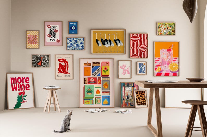 Pode incluir: Uma cole&ccedil;&atilde;o de impress&otilde;es de arte emolduradas em v&aacute;rios tamanhos e estilos, incluindo designs abstratos, ilustra&ccedil;&otilde;es e tipografia. A paleta de cores inclui vermelho, amarelo, azul e rosa. Um gato est&aacute; sentado no ch&atilde;o em frente &agrave;s obras de arte de parede.