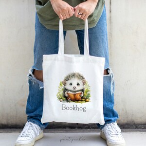 May include: White tote bag with a cartoon hedgehog reading a book. The bag has the text "Bookhog" printed on it.