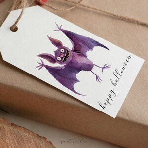 Puede incluir: Una etiqueta de regalo blanca con un murci&eacute;lago de dibujos animados morado con ojos grandes y el texto "happy halloween" en escritura cursiva negra.