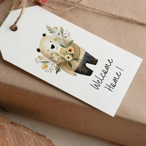 Peut inclure: Une &eacute;tiquette cadeau blanche avec un ours brun tenant un bouquet de fleurs jaunes et blanches. Le texte "Welcome Home!" est &eacute;crit en cursive noire sur l'&eacute;tiquette.