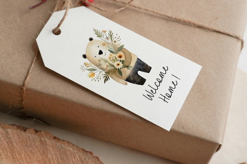 Peut inclure: Une &eacute;tiquette cadeau blanche avec un ours brun tenant un bouquet de fleurs jaunes et blanches. Le texte "Welcome Home!" est &eacute;crit en cursive noire sur l'&eacute;tiquette.