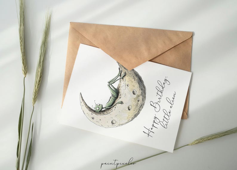 Puede incluir: Una tarjeta blanca con un extraterrestre verde sentado en una luna creciente. El texto en la tarjeta dice "Feliz cumplea&ntilde;os, peque&ntilde;o extraterrestre."