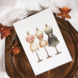 May include: A watercolor illustration of three vintage dress forms with floral details. The dress forms are wearing different styles of dresses, including a black lace dress, a beige dress with floral details, and a white dress with floral details.