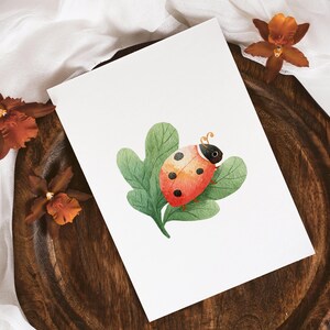 May include: Watercolor illustration of a ladybug on a leaf. The ladybug is red with black spots and sits on a green leaf. The artwork is on a white card, with brown orchids and a wooden tray.