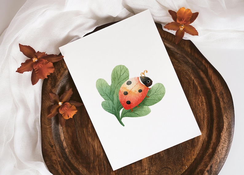 May include: Watercolor illustration of a ladybug on a leaf. The ladybug is red with black spots and sits on a green leaf. The artwork is on a white card, with brown orchids and a wooden tray.