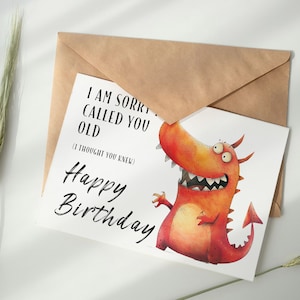 May include: A white card with a red and orange cartoon dragon with three eyes and a toothy grin. The card says "I am sorry I called you old (I thought you knew) Happy Birthday".
