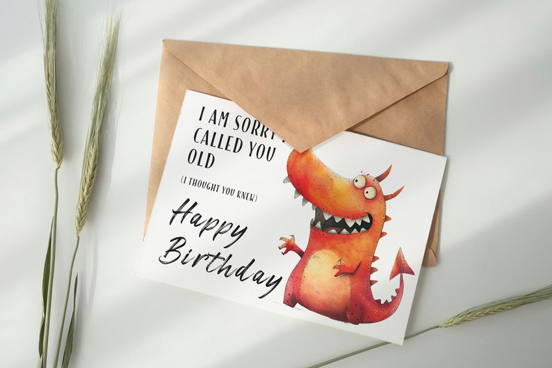 May include: A white card with a red and orange cartoon dragon with three eyes and a toothy grin. The card says "I am sorry I called you old (I thought you knew) Happy Birthday".