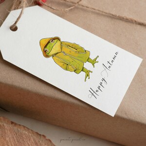May include: A white gift tag with a green frog wearing a yellow raincoat. The tag has the text "Happy Autumn" written in black cursive.