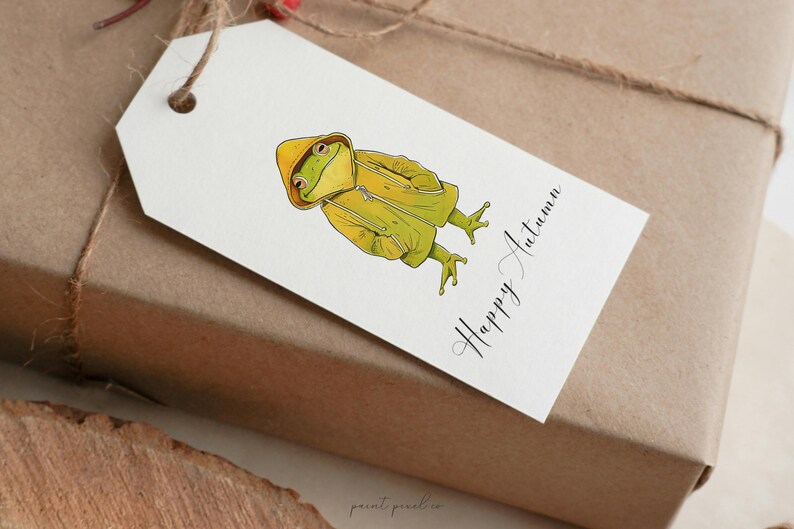 May include: A white gift tag with a green frog wearing a yellow raincoat. The tag has the text "Happy Autumn" written in black cursive.