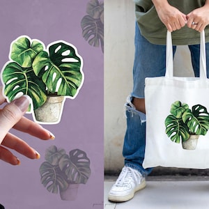 May include: A white tote bag with a green watercolor illustration of a potted monstera plant. The bag has a long handle and is made of canvas.