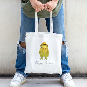 May include: White tote bag with a green frog wearing a yellow raincoat and the text "Happy Autumn".