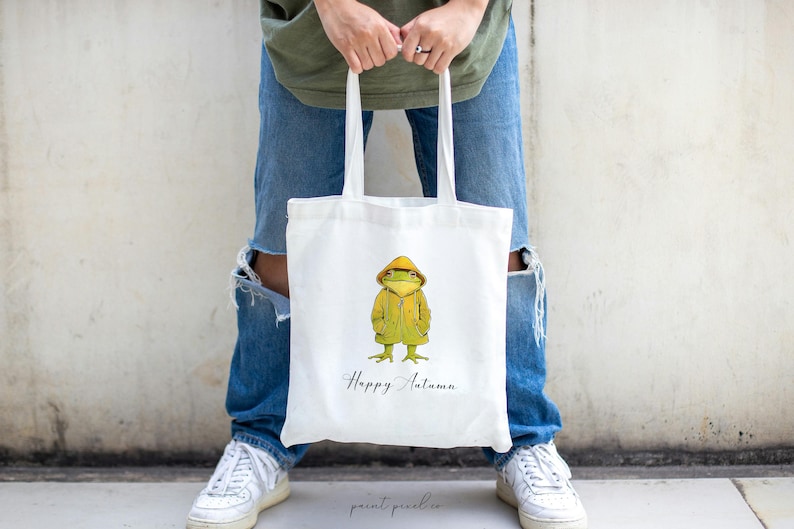 May include: White tote bag with a green frog wearing a yellow raincoat and the text "Happy Autumn".