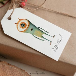 Puede incluir: Una etiqueta de regalo blanca con una ilustraci&oacute;n de dibujos animados de un monstruo azul y verde con un ojo grande. El texto "hello there!" est&aacute; escrito en cursiva negra en la etiqueta.