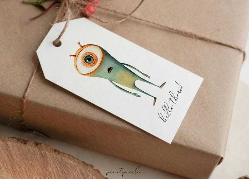 Puede incluir: Una etiqueta de regalo blanca con una ilustraci&oacute;n de dibujos animados de un monstruo azul y verde con un ojo grande. El texto "hello there!" est&aacute; escrito en cursiva negra en la etiqueta.