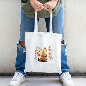 Peut inclure: Un sac fourre-tout en toile blanche avec une illustration mignonne d'un renard assis dans un champ de fleurs orange. Le renard est brun et blanc avec une queue touffue.