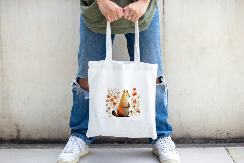 Peut inclure: Un sac fourre-tout en toile blanche avec une illustration mignonne d'un renard assis dans un champ de fleurs orange. Le renard est brun et blanc avec une queue touffue.