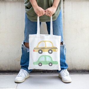 May include: A white tote bag with a yellow and green car design. The bag has two handles and is being held by a person.