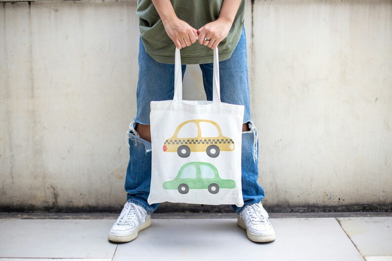 May include: A white tote bag with a yellow and green car design. The bag has two handles and is being held by a person.