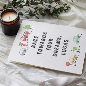 May include: A white card with the text "Race towards your dreams, Lucas."  The card is decorated with a red fire truck, a yellow car, a green car, and a stop sign. The card is on a white fabric surface.
