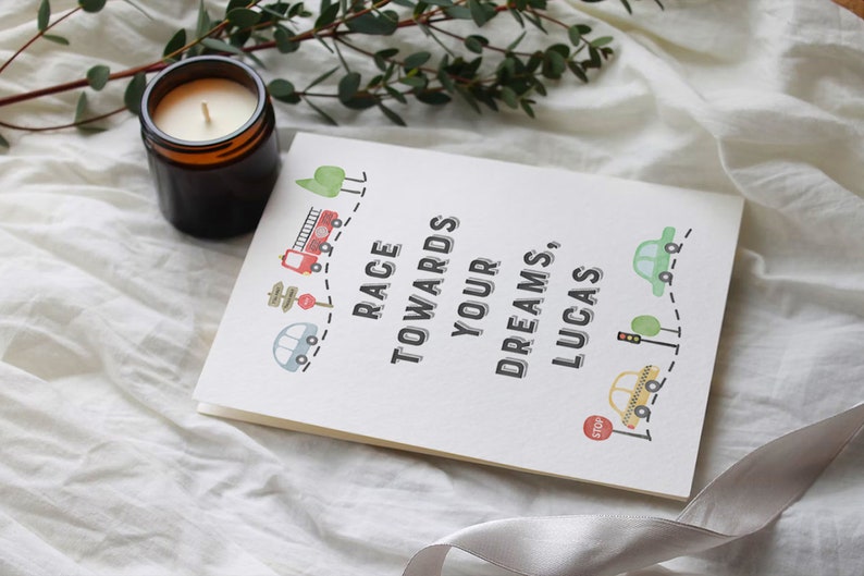 May include: A white card with the text "Race towards your dreams, Lucas."  The card is decorated with a red fire truck, a yellow car, a green car, and a stop sign. The card is on a white fabric surface.