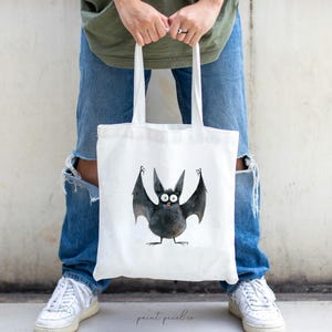 Puede incluir: Una bolsa de tela blanca con una ilustraci&oacute;n de un murci&eacute;lago negro. El murci&eacute;lago tiene ojos grandes y est&aacute; sosteniendo sus alas hacia arriba. La bolsa tiene un asa negra.