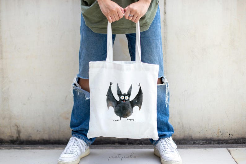 Puede incluir: Una bolsa de tela blanca con una ilustraci&oacute;n de un murci&eacute;lago negro. El murci&eacute;lago tiene ojos grandes y est&aacute; sosteniendo sus alas hacia arriba. La bolsa tiene un asa negra.