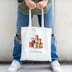May include: White tote bag with a colorful illustration of spools of thread and the text "love for sewing".
