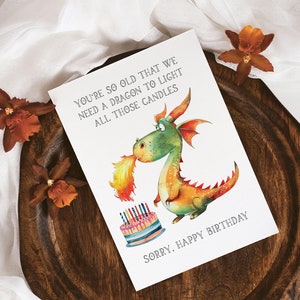 May include: A humorous birthday card featuring a cartoon dragon blowing fire on a birthday cake with candles. The card reads "You're so old that we need a dragon to light all those candles. Sorry, Happy Birthday."
