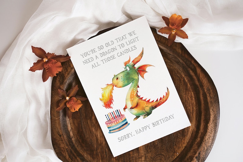 May include: A humorous birthday card featuring a cartoon dragon blowing fire on a birthday cake with candles. The card reads "You're so old that we need a dragon to light all those candles. Sorry, Happy Birthday."