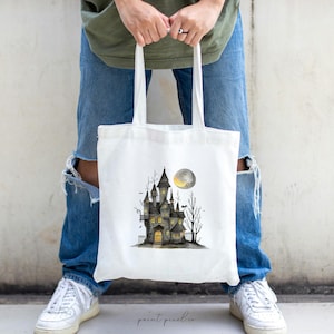 May include: A white tote bag with a black and white illustration of a haunted house with a full moon. The house has windows and a door with a light shining from inside.