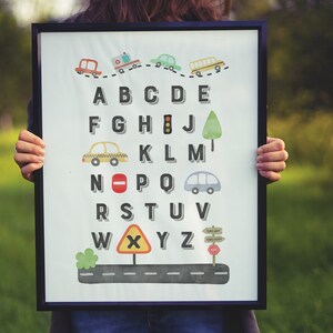 May include: A white framed alphabet chart with a road theme. The letters are in black and are surrounded by various road signs and vehicles, including a school bus, ambulance, taxi, and stop sign. The letters are A-Z.