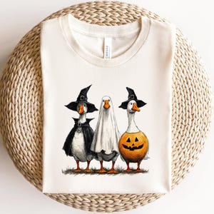May include: Off-white t-shirt featuring three geese dressed for Halloween. One goose wears a witch hat and cape, another is in a ghost costume, and the third is a jack-o'-lantern. The geese are standing on grass.