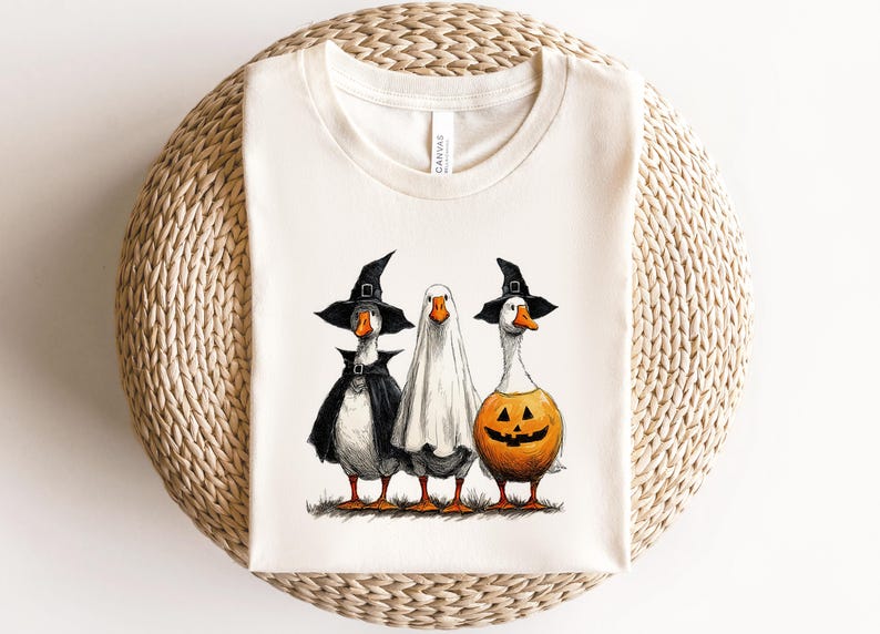 May include: Off-white t-shirt featuring three geese dressed for Halloween. One goose wears a witch hat and cape, another is in a ghost costume, and the third is a jack-o'-lantern. The geese are standing on grass.