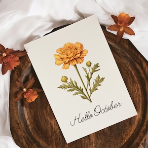 Op de afbeelding: Een witte kaart met een handgetekende illustratie van een gele bloem met groene bladeren en de tekst "Hello October". De kaart ligt op een houten oppervlak met een witte stof op de achtergrond.