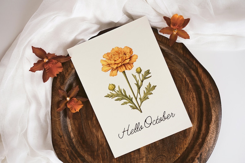 Op de afbeelding: Een witte kaart met een handgetekende illustratie van een gele bloem met groene bladeren en de tekst "Hello October". De kaart ligt op een houten oppervlak met een witte stof op de achtergrond.