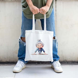 May include: A white tote bag with a cartoon illustration of an older woman doing a yoga pose. The bag has two handles and is being held by a person wearing blue jeans and white sneakers.
