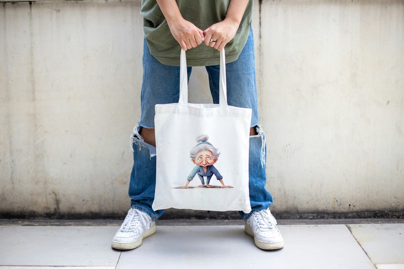 May include: A white tote bag with a cartoon illustration of an older woman doing a yoga pose. The bag has two handles and is being held by a person wearing blue jeans and white sneakers.