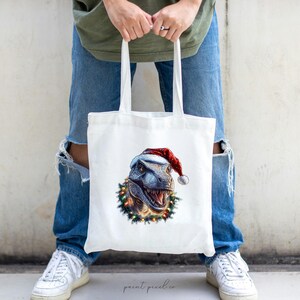 May include: White tote bag with a Christmas-themed design featuring a dinosaur wearing a Santa hat and surrounded by a wreath of holly and lights.