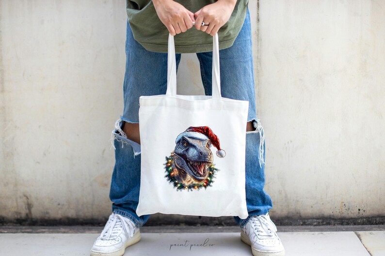May include: White tote bag with a Christmas-themed design featuring a dinosaur wearing a Santa hat and surrounded by a wreath of holly and lights.