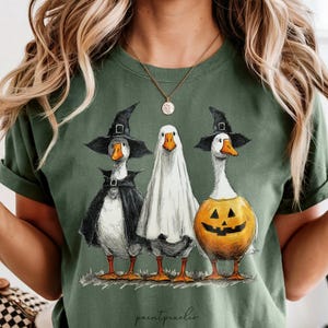 May include: Green t-shirt featuring three geese dressed in Halloween costumes. One goose wears a witch hat and cape, another is a ghost, and the third is a jack-o'-lantern. The words "paintpeaches" are at the bottom.