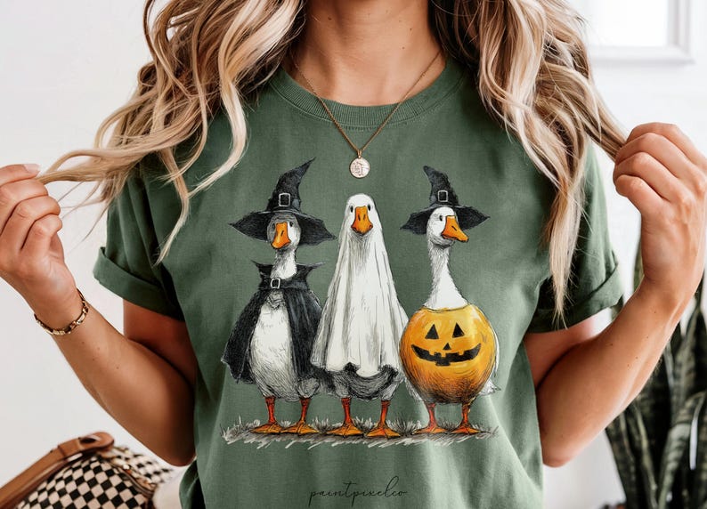 May include: Green t-shirt featuring three geese dressed in Halloween costumes. One goose wears a witch hat and cape, another is a ghost, and the third is a jack-o'-lantern. The words "paintpeaches" are at the bottom.