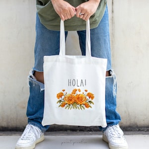 May include: A white tote bag with the word "HOLAI" printed in black and a floral design featuring orange marigolds.