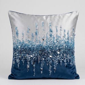 Può includere: Un cuscino quadrato decorativo con una parte superiore argentata e una parte inferiore in velluto blu scuro. Il cuscino è impreziosito da elementi a cascata simili a cristalli blu e argento, creando un effetto scintillante. Il cuscino ha un bordo blu scuro.