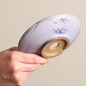 May include: A handmade ceramic dish with a light purple glaze and a brown base. The dish has a circular shape with a hole in the center of the base. The glaze has a speckled appearance with blue accents.